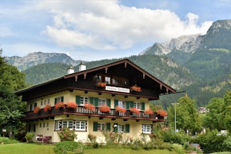 A picturesque chalet-style building with wooden balconies decorated with red flowers, surrounded by lush greenery and set against a backdrop of towering mountains under a partly cloudy sky.