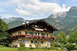 A picturesque chalet-style building with wooden balconies decorated with red flowers, surrounded by lush greenery and set against a backdrop of towering mountains under a partly cloudy sky.