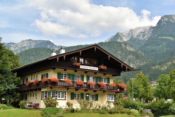 A picturesque chalet-style building with wooden balconies decorated with red flowers, surrounded by lush greenery and set against a backdrop of towering mountains under a partly cloudy sky.