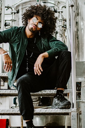 A person with an afro hairstyle and sunglasses sits on a metal staircase, wearing a dark green jacket, black pants, and black sneakers. The setting appears industrial, with metallic pipes and equipment in the background.