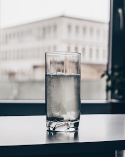 A glass of water with ice cubes sits on a table near a window, with an out-of-focus building visible in the background.