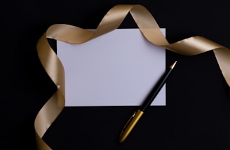 A blank white card surrounded by a luxurious gold ribbon on a dark background. A black and gold pen is placed beside the card, adding a touch of elegance.