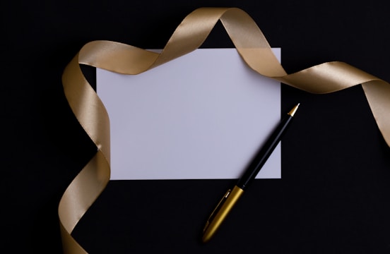 A blank white card surrounded by a luxurious gold ribbon on a dark background. A black and gold pen is placed beside the card, adding a touch of elegance.