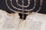 An ancient manuscript with Hebrew text is set beneath a metal menorah. The pages of the manuscript are aged and torn, indicating its historical significance.