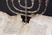 An ancient manuscript with Hebrew text is set beneath a metal menorah. The pages of the manuscript are aged and torn, indicating its historical significance.