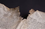 A close-up of an ancient, torn parchment displaying handwritten text in Hebrew script. The edges of the parchment are frayed and browned, suggesting age and wear. The text is in black ink against a light yellowed background, with the tear creating a dramatic line through the middle.