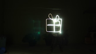 A long-exposure photograph captures light trails forming the shape of a gift box against a dark background. The light creates the outline of a box with a bow on top, illuminating the surroundings subtly.