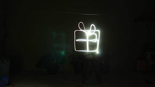 A long-exposure photograph captures light trails forming the shape of a gift box against a dark background. The light creates the outline of a box with a bow on top, illuminating the surroundings subtly.