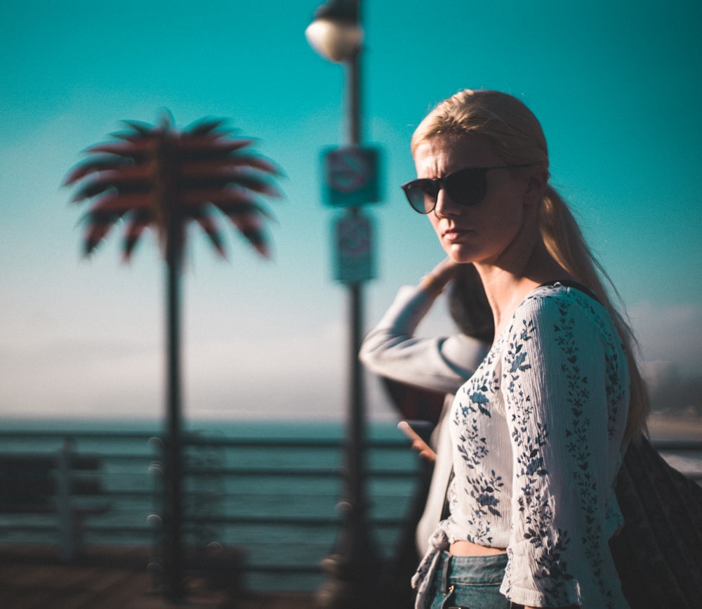 woman in white floral lace shirt and blue denim jacket wearing black sunglasses standing near palm