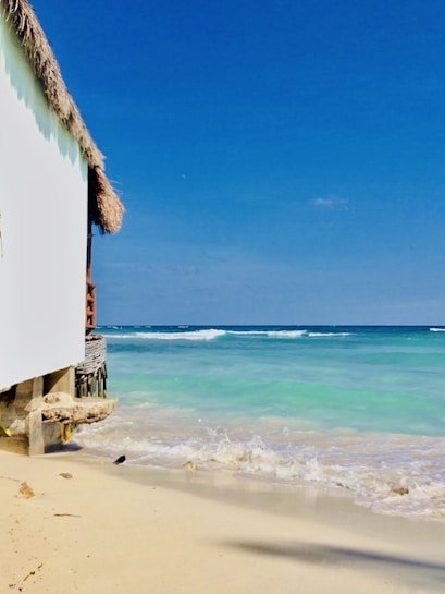 A serene beach scene with turquoise waters gently lapping against a sandy shore. A structure with a thatched roof and white walls borders the left side, while the vibrant blue sky stretches above.
