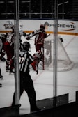 A hockey game scene with players in action near a goalpost. The players are wearing red and white uniforms with protective gear, including helmets. An official wearing a black and white striped uniform is present, observing the play closely. There is a hockey net visible in the background with advertisements on the arena walls.