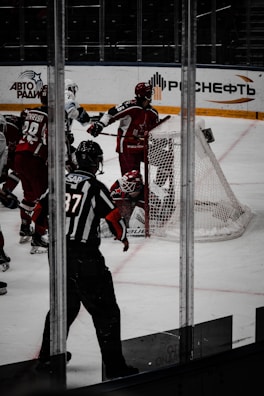 A hockey game scene with players in action near a goalpost. The players are wearing red and white uniforms with protective gear, including helmets. An official wearing a black and white striped uniform is present, observing the play closely. There is a hockey net visible in the background with advertisements on the arena walls.