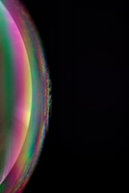 A vibrant and colorful spherical object displays a gradient of rainbow hues against a dark background. The surface appears glossy and reflective, showing a blend of greens, pinks, and purples with softer transitions at the edges.