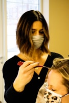 A person with dark shoulder-length hair and a face mask is applying a treatment or dye to another person's hair using a brush. The second person has light-colored hair and is wearing a face mask with a leopard print. They are situated in a bright room with a large window in the background.