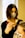 A person with dark shoulder-length hair and a face mask is applying a treatment or dye to another person's hair using a brush. The second person has light-colored hair and is wearing a face mask with a leopard print. They are situated in a bright room with a large window in the background.