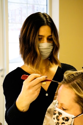 A person with dark shoulder-length hair and a face mask is applying a treatment or dye to another person's hair using a brush. The second person has light-colored hair and is wearing a face mask with a leopard print. They are situated in a bright room with a large window in the background.