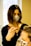 A person with dark shoulder-length hair and a face mask is applying a treatment or dye to another person's hair using a brush. The second person has light-colored hair and is wearing a face mask with a leopard print. They are situated in a bright room with a large window in the background.