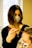 A person with dark shoulder-length hair and a face mask is applying a treatment or dye to another person's hair using a brush. The second person has light-colored hair and is wearing a face mask with a leopard print. They are situated in a bright room with a large window in the background.