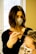 A person with dark shoulder-length hair and a face mask is applying a treatment or dye to another person's hair using a brush. The second person has light-colored hair and is wearing a face mask with a leopard print. They are situated in a bright room with a large window in the background.