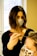 A person with dark shoulder-length hair and a face mask is applying a treatment or dye to another person's hair using a brush. The second person has light-colored hair and is wearing a face mask with a leopard print. They are situated in a bright room with a large window in the background.