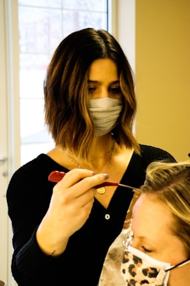 A person with dark shoulder-length hair and a face mask is applying a treatment or dye to another person's hair using a brush. The second person has light-colored hair and is wearing a face mask with a leopard print. They are situated in a bright room with a large window in the background.