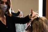 A hairstylist wearing a face mask is working on a client's hair, which is covered with aluminum foils for coloring. The client, also wearing a face mask with a leopard print, is looking at the mirror. The scene is brightly lit by a vertical light strip next to the mirror, creating a focused working environment.