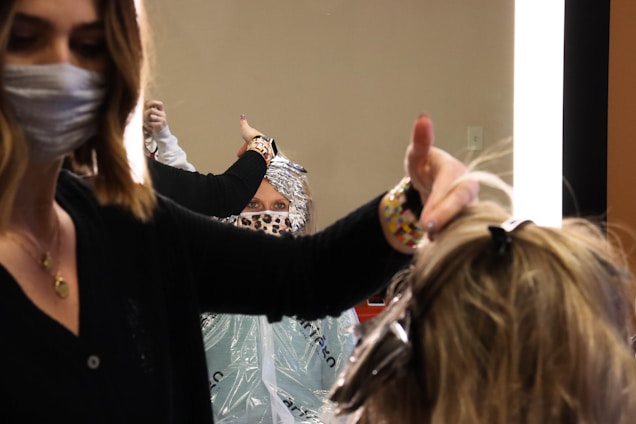 A hairstylist wearing a face mask is working on a client's hair, which is covered with aluminum foils for coloring. The client, also wearing a face mask with a leopard print, is looking at the mirror. The scene is brightly lit by a vertical light strip next to the mirror, creating a focused working environment.