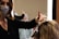 A hairstylist wearing a face mask is working on a client's hair, which is covered with aluminum foils for coloring. The client, also wearing a face mask with a leopard print, is looking at the mirror. The scene is brightly lit by a vertical light strip next to the mirror, creating a focused working environment.