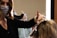 A hairstylist wearing a face mask is working on a client's hair, which is covered with aluminum foils for coloring. The client, also wearing a face mask with a leopard print, is looking at the mirror. The scene is brightly lit by a vertical light strip next to the mirror, creating a focused working environment.
