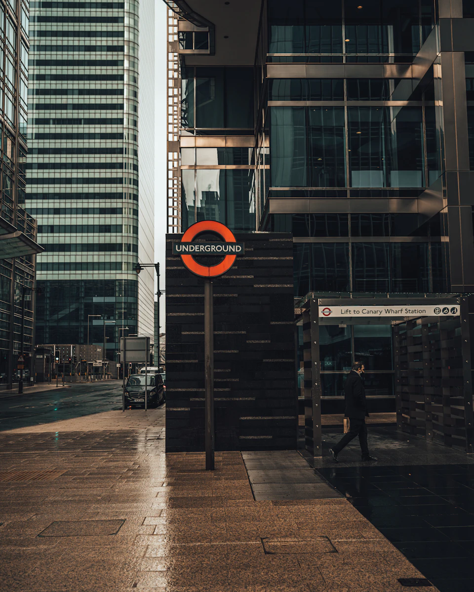 The image depicts a bustling city scene, with tall buildings lining the street and a subway station visible on the right side. The station's entrance is marked by an orange sign that reads "Underground", indicating its location within the underground system. A man can be seen walking towards the station, adding a sense of movement to the urban landscape.

On the left side of the image, there are several cars parked or driving along the street, contributing to the busy atmosphere. The sky overhead is overcast, casting a soft light on the scene and creating an interesting interplay between shadows and highlights.