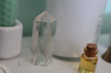 A clear quartz crystal stands upright on a surface next to a small bottle filled with yellow liquid, sealed with a cork. Behind them is a large white candle and a green textured candle in a white holder.