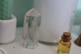 A clear quartz crystal stands upright on a surface next to a small bottle filled with yellow liquid, sealed with a cork. Behind them is a large white candle and a green textured candle in a white holder.