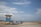 A small boat named Karibana rests on the sandy shore next to a simple, sky-blue lifeguard hut. The backdrop features a tranquil seascape with calm waters and a partly cloudy sky. Large rocks separate the sandy beach from the water.
