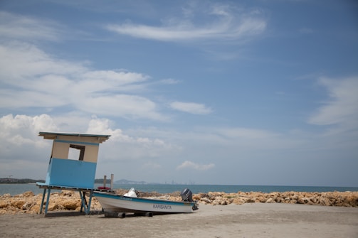 A small boat named Karibana rests on the sandy shore next to a simple, sky-blue lifeguard hut. The backdrop features a tranquil seascape with calm waters and a partly cloudy sky. Large rocks separate the sandy beach from the water.