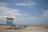 A small boat named Karibana rests on the sandy shore next to a simple, sky-blue lifeguard hut. The backdrop features a tranquil seascape with calm waters and a partly cloudy sky. Large rocks separate the sandy beach from the water.