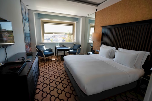 A clean and modern hotel room with a large bed covered in white linens. There is a decorative carpet with a geometric pattern on the floor. Near the window, there are two upholstered chairs and a small table with two bottles of water. A flat-screen TV is mounted on the wall opposite the bed. The room features neutral colors and ample natural light from the window overlooking a cityscape.