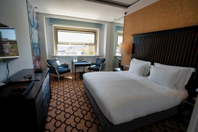 A clean and modern hotel room with a large bed covered in white linens. There is a decorative carpet with a geometric pattern on the floor. Near the window, there are two upholstered chairs and a small table with two bottles of water. A flat-screen TV is mounted on the wall opposite the bed. The room features neutral colors and ample natural light from the window overlooking a cityscape.