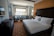 A clean and modern hotel room with a large bed covered in white linens. There is a decorative carpet with a geometric pattern on the floor. Near the window, there are two upholstered chairs and a small table with two bottles of water. A flat-screen TV is mounted on the wall opposite the bed. The room features neutral colors and ample natural light from the window overlooking a cityscape.