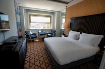 A clean and modern hotel room with a large bed covered in white linens. There is a decorative carpet with a geometric pattern on the floor. Near the window, there are two upholstered chairs and a small table with two bottles of water. A flat-screen TV is mounted on the wall opposite the bed. The room features neutral colors and ample natural light from the window overlooking a cityscape.