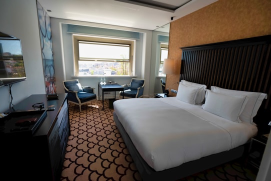 A clean and modern hotel room with a large bed covered in white linens. There is a decorative carpet with a geometric pattern on the floor. Near the window, there are two upholstered chairs and a small table with two bottles of water. A flat-screen TV is mounted on the wall opposite the bed. The room features neutral colors and ample natural light from the window overlooking a cityscape.