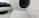 A close-up view of a white, spherical rotating webcam or security camera, placed on a smooth surface. The lens is slightly tilted upwards, with a black center surrounded by a white casing. There is a portion of a rectangular black object in the background.