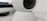 A close-up view of a white, spherical rotating webcam or security camera, placed on a smooth surface. The lens is slightly tilted upwards, with a black center surrounded by a white casing. There is a portion of a rectangular black object in the background.