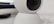 A close-up view of a white, spherical rotating webcam or security camera, placed on a smooth surface. The lens is slightly tilted upwards, with a black center surrounded by a white casing. There is a portion of a rectangular black object in the background.