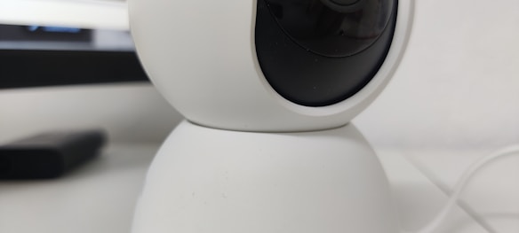 A close-up view of a white, spherical rotating webcam or security camera, placed on a smooth surface. The lens is slightly tilted upwards, with a black center surrounded by a white casing. There is a portion of a rectangular black object in the background.