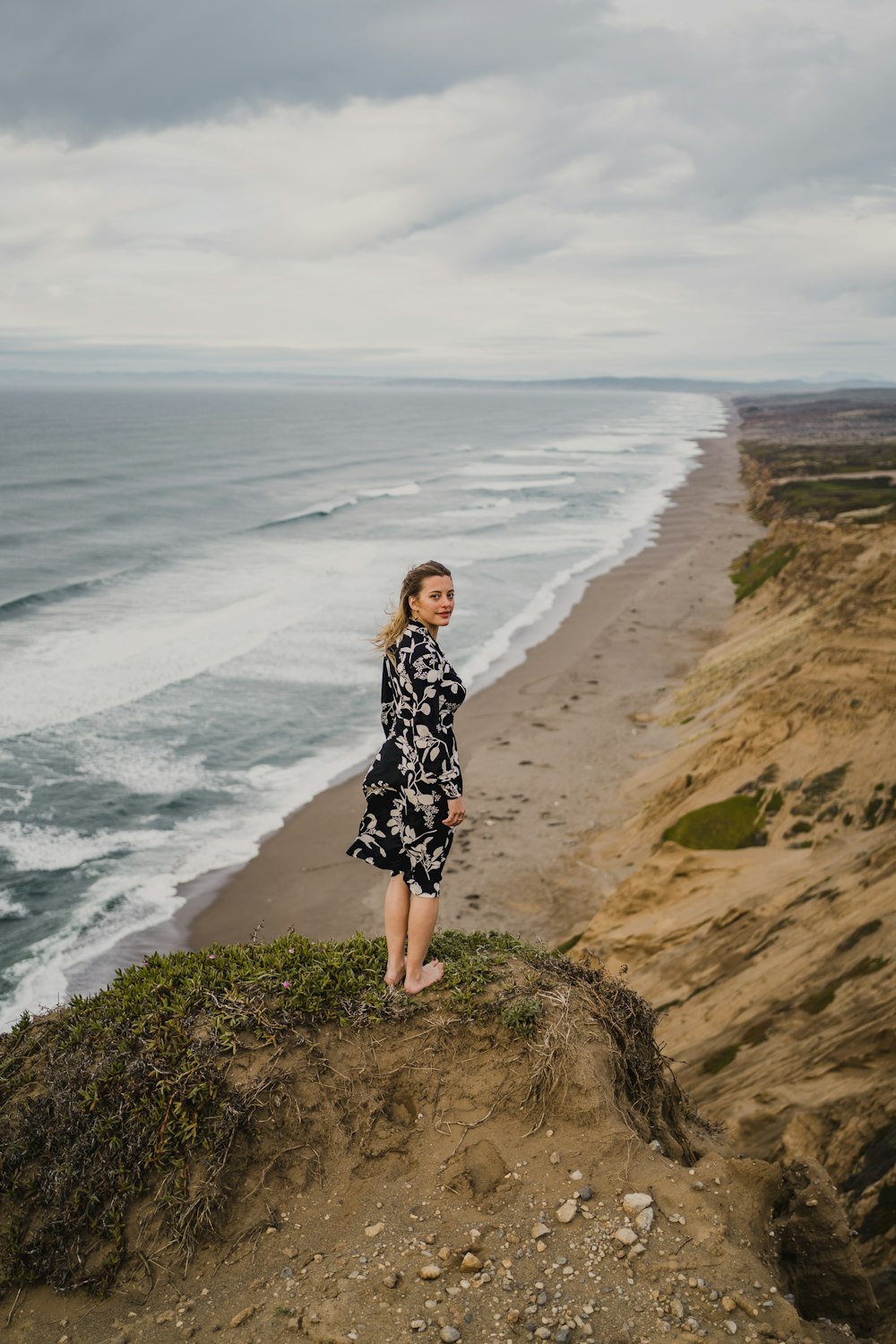 woman in black and white floral dress standing on brown rock near body of water during