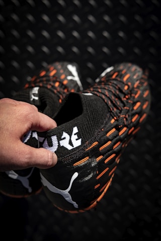 A hand holding a pair of black sports shoes featuring a white logo and the word 'FUTURE' in bold letters. The shoes have orange accents and textured patterns on a dark, patterned background.