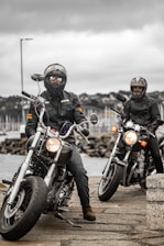 Two motorcyclists wearing helmets and leather jackets are posed on their bikes near a waterfront on a cloudy day. In the background, a marina with numerous sailboats is visible under an overcast sky.