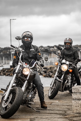 Two motorcyclists wearing helmets and leather jackets are posed on their bikes near a waterfront on a cloudy day. In the background, a marina with numerous sailboats is visible under an overcast sky.