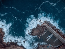 An aerial view of a coastal harbor with waves crashing against the breakwater. The image captures a few small boats docked in the marina, surrounded by the deep blue of the ocean. The rocky shoreline and man-made structures such as piers and barriers are visible.