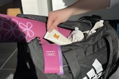 A hand holding a tube of muscle relief cream is reaching into a black sports bag. Inside the bag, there is a pink rolled-up yoga mat with floral patterns and a purple heavy resistance band. The bag also contains white items, possibly clothing.